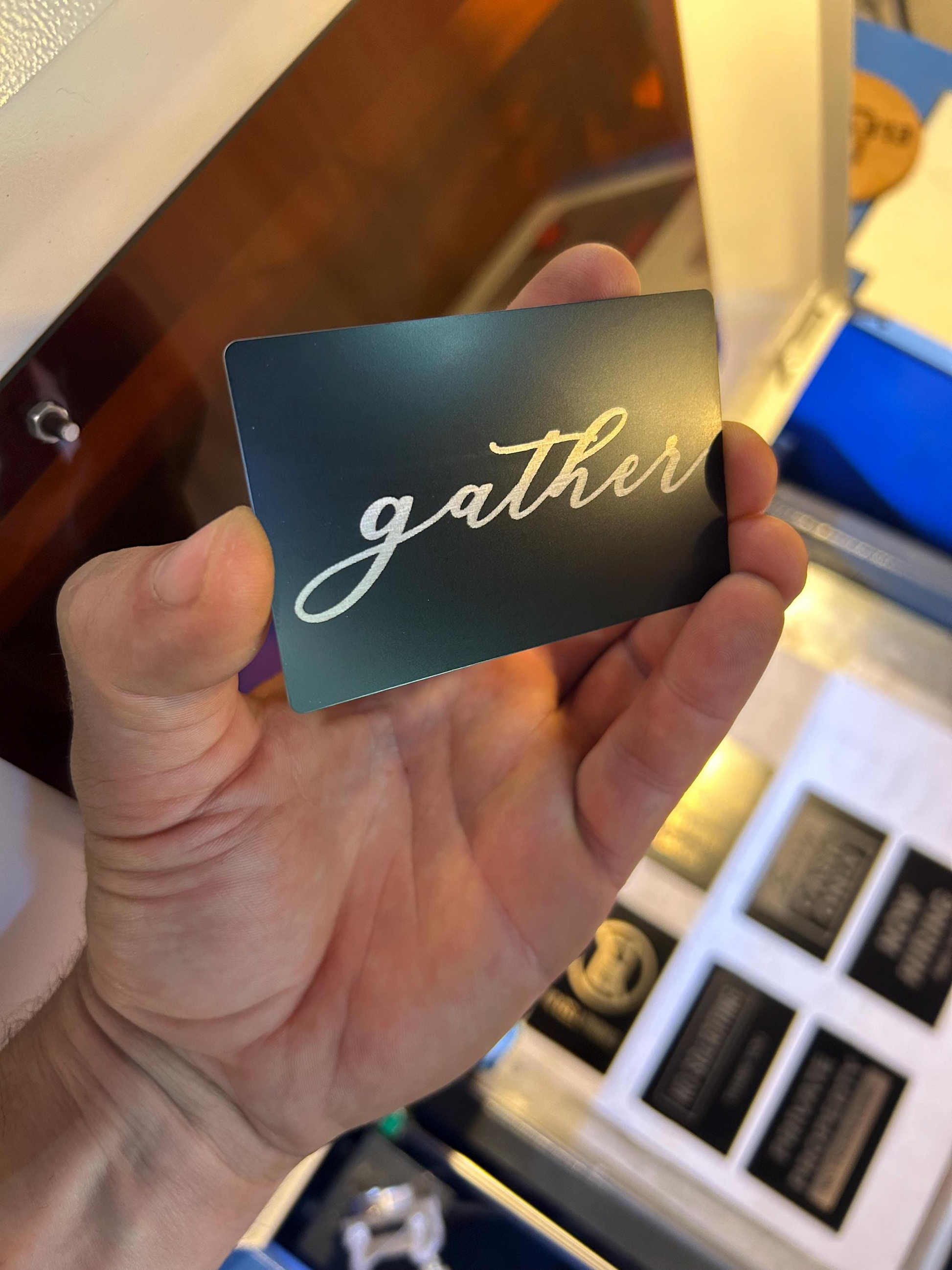 Hand holding a black card with 'gather' engraving in silver cursive text, blurred background