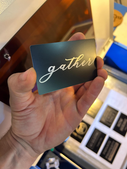 Hand holding a black card with 'gather' engraving in silver cursive text, blurred background