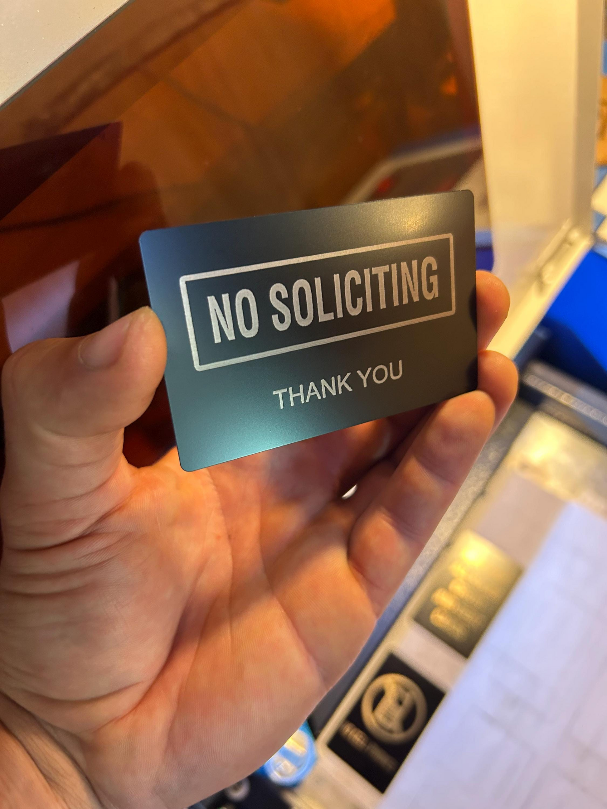 Person holding a metallic black and silver engraved signage card featuring “No Soliciting” text