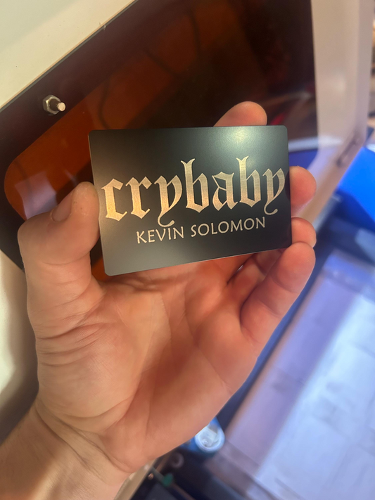 Hand holding a 'crybaby' card with  personalized engraving details, blurred machine background
