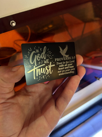 Hand holding a black/silver metal card with 'In God We Trust' and Proverbs 3:5 text.