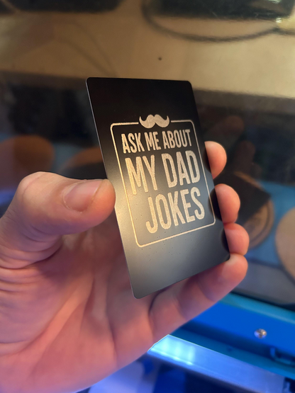 Detailed shot of laser cut novelty gift card – “Ask Me About My Dad Jokes” engraved in silver on black background with mustache graphic