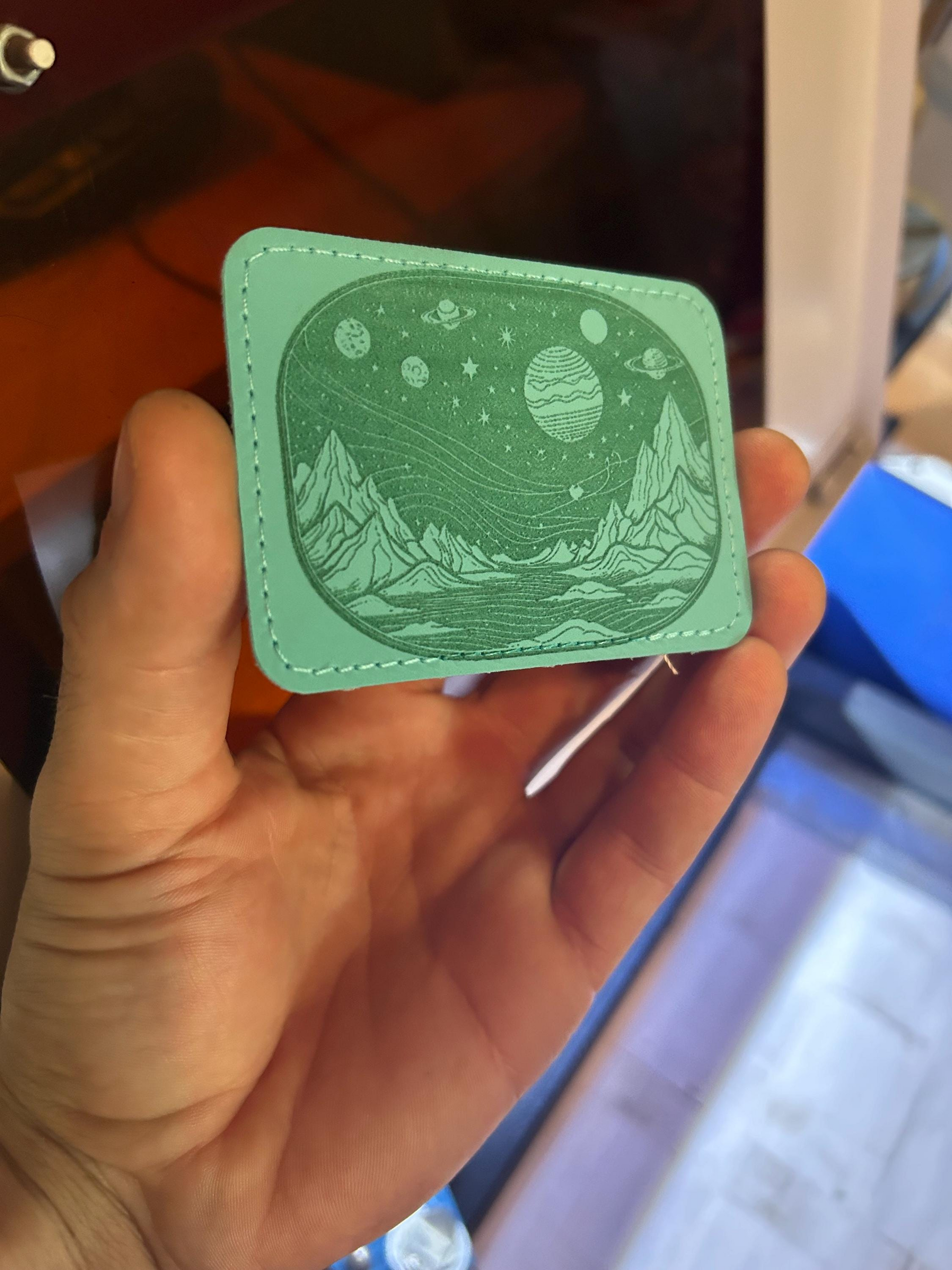 Hand holding a teal 3.5 x 2.5" leather patch with space planet stars galaxy mountain design