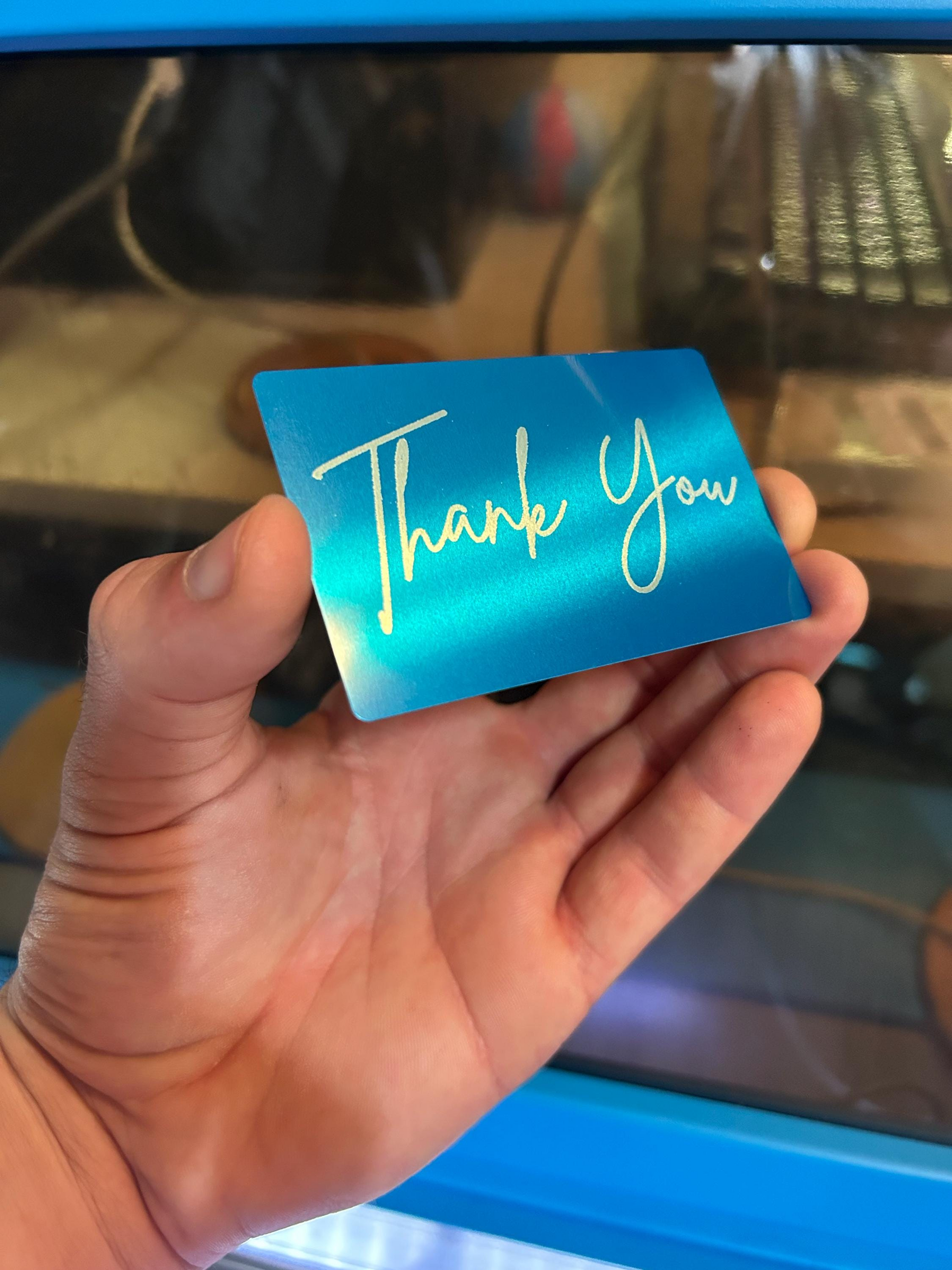 Hand holding a blue and silver metal greeting card with 'Thank You' text against a blurred background