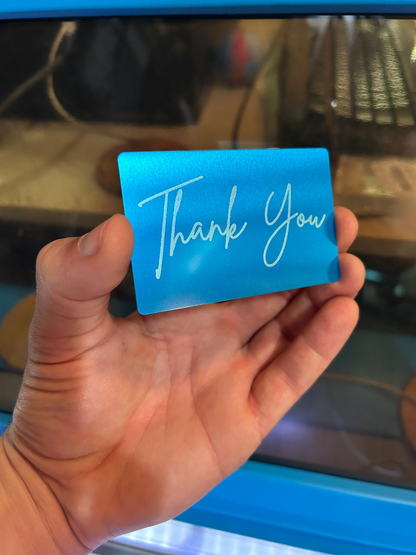Hand holding a blue and silver metal gift card with 'Thank You' laser engraved on it, blurred background
