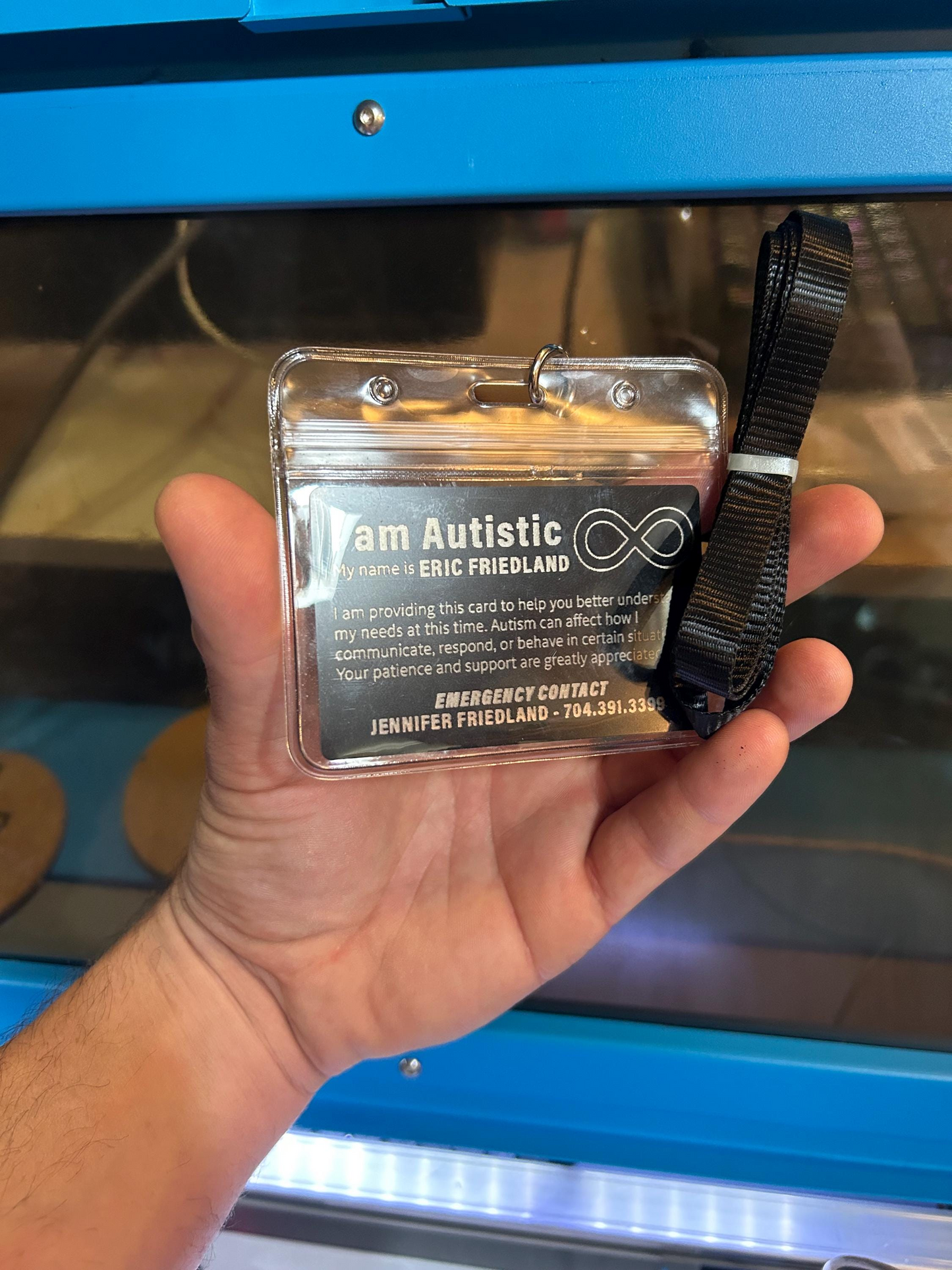 Hand holding a cardin a lanyard badge pass holder with personalized autism awareness text and a black strap in front of a blue machine.