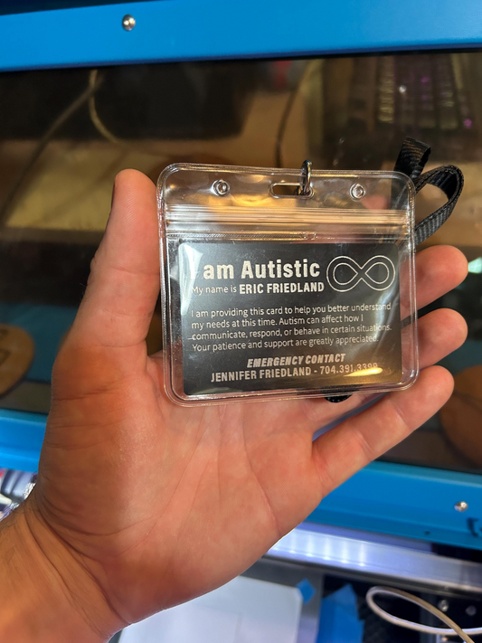 Hand holding a clear card holder and lanyard, with laser engraved personalized text about autism in front of a blue machine.