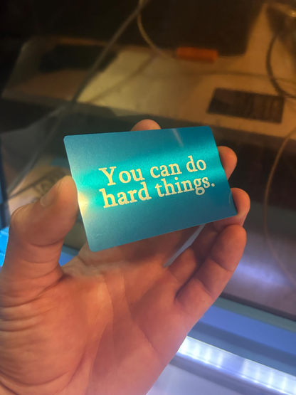 Hand holding a blue silver/blue metal keepsake card with 'You can do hard things' text  in anodized aluminum.