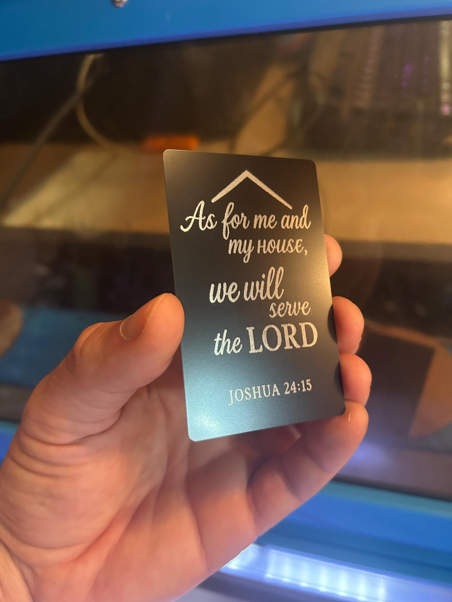 Hand holding a black and silver laser engraved metal keepsake card with religious inspirational text against a blurred background