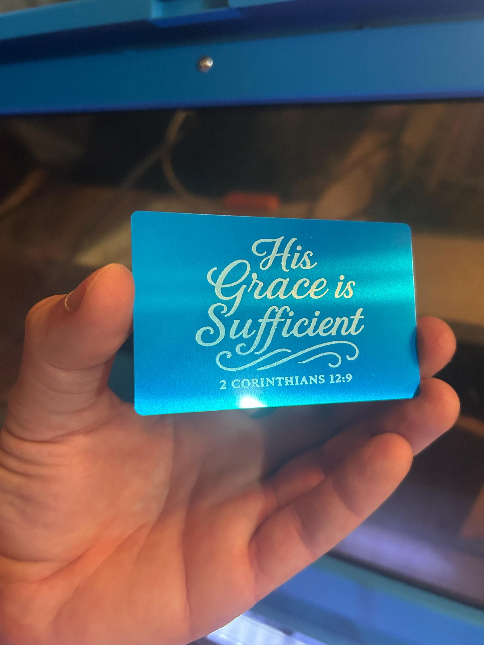 Blue metal keepsake placard with 'His Grace is Sufficient' text held by a hand