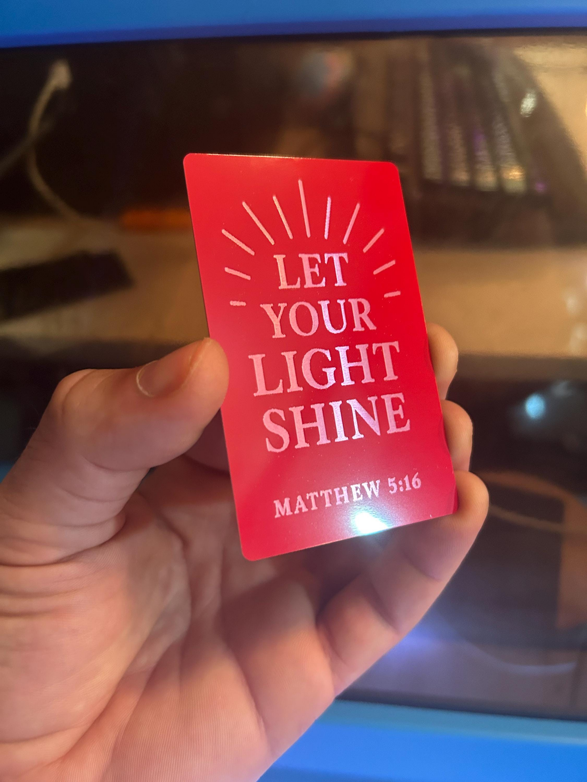 Hand holding a red and silver metal keepsake card engraved with “Let Your Light Shine” and Matthew 5:16 against blurred laser machine background