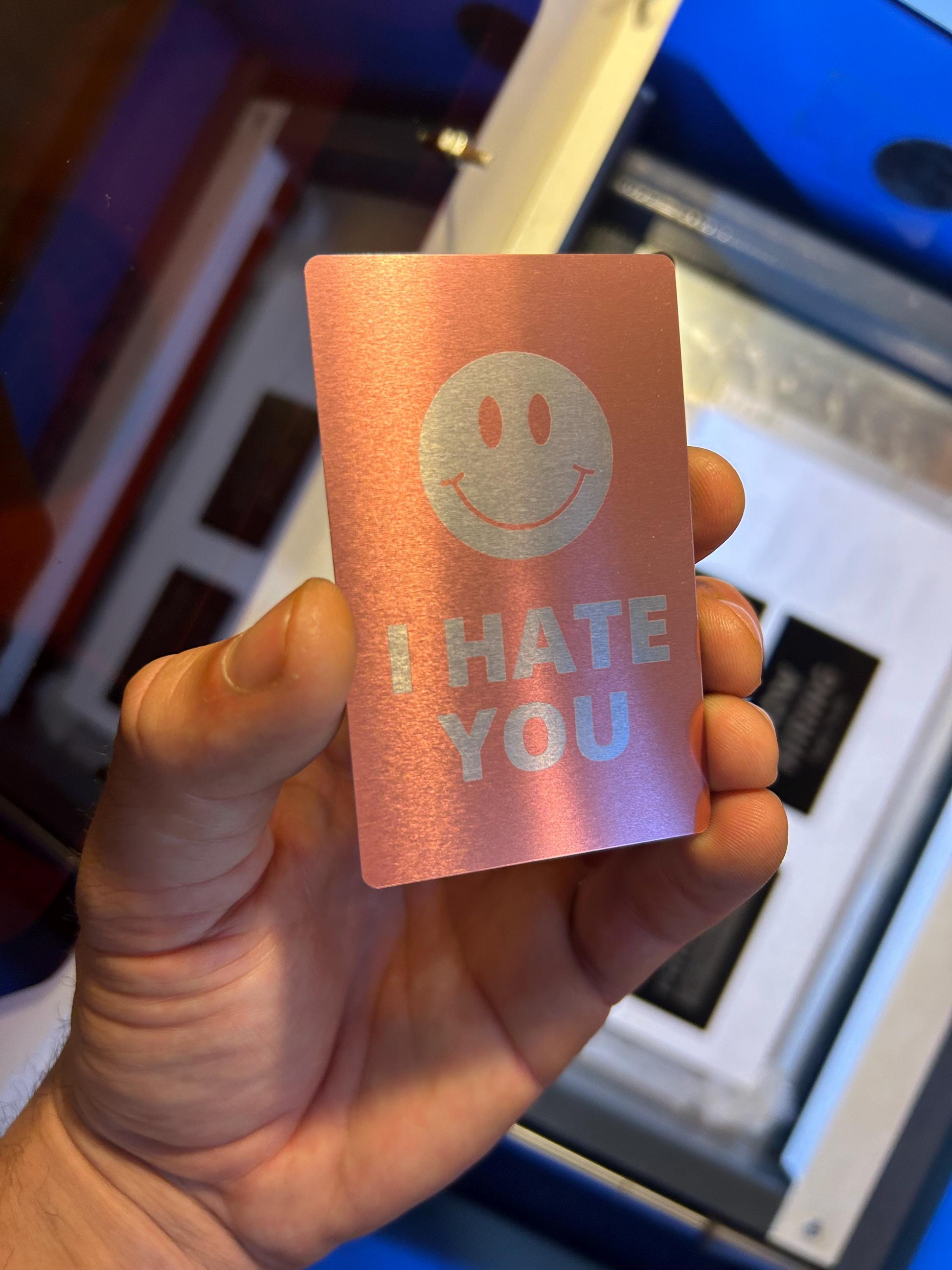 Hand holding an anodized aluminum gag gift card with a smiley face and 'I HATE YOU' text against a blurred indoor background.