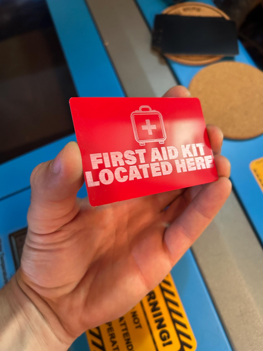 Red metal card with 'First Aid Kit Located Here' text held by a hand against a blurred background
