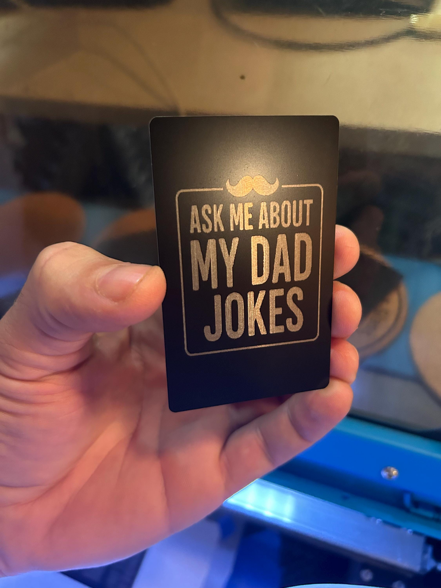 Angled shot of custom black/silver metal joke card featuring “Ask Me About My Dad Jokes” and engraved mustache artwork