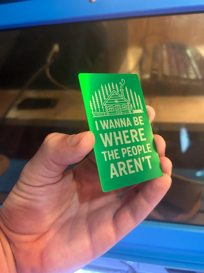 Hand holding a metal keepsake card with "I wanna be where the people aren't" typography text and cabin illustration, photographed in front of a blue machine