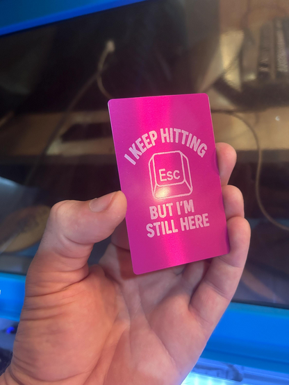 Hand holding a laser engraved metal keepsake card with I Keep Hitting Escape, But I'm Still Here text and graphic artwork on a purple/silver anodized aluminum card