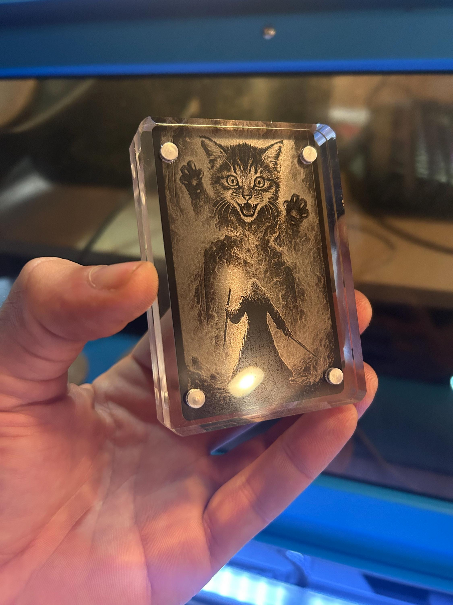 Hand holding an acrylic display case with a engraved design of a gigantic cat and adventurer mage illustration, against a blurred background.