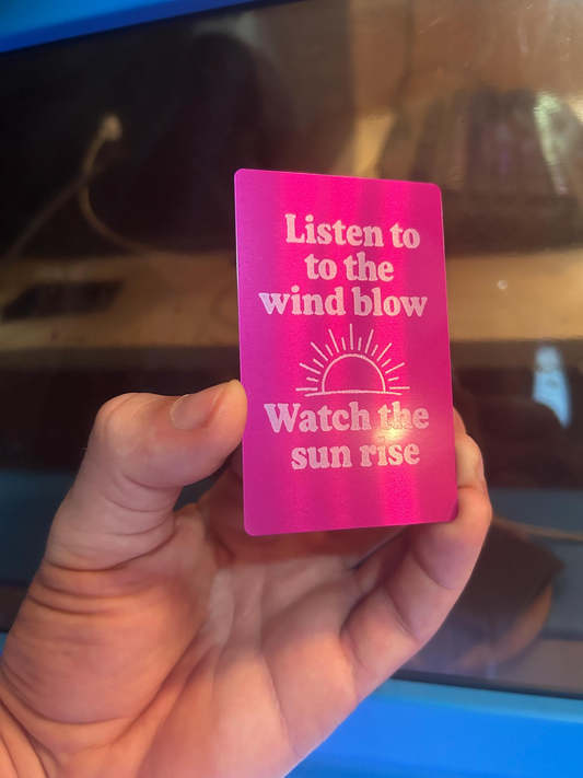 Hand holding a purple/silver metal gift card with text and a sun illustration, against a blurred machine background.
