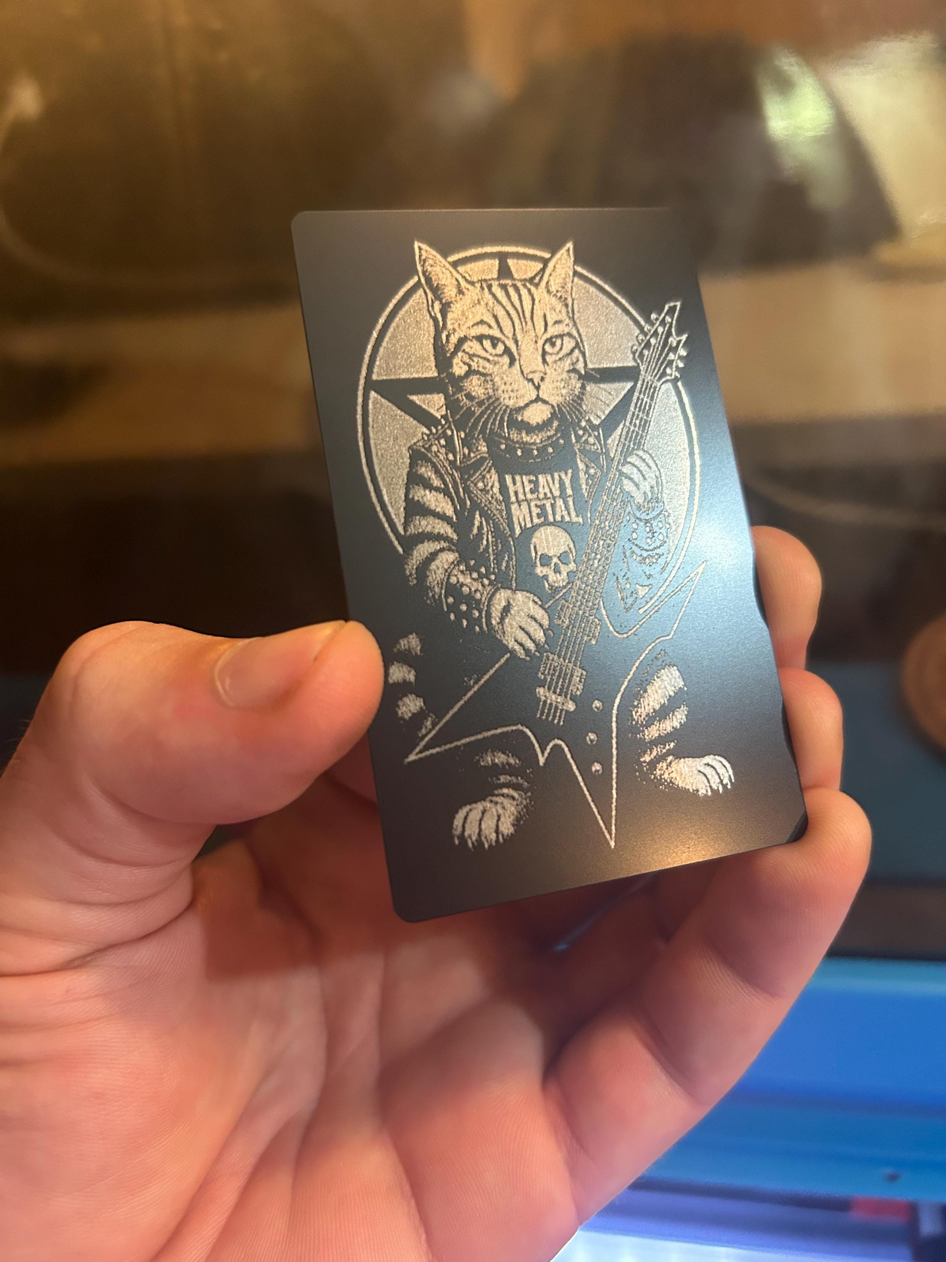 Hand holding a black and silver metal laser engraved keepsake card with a kitten playing guitar design.