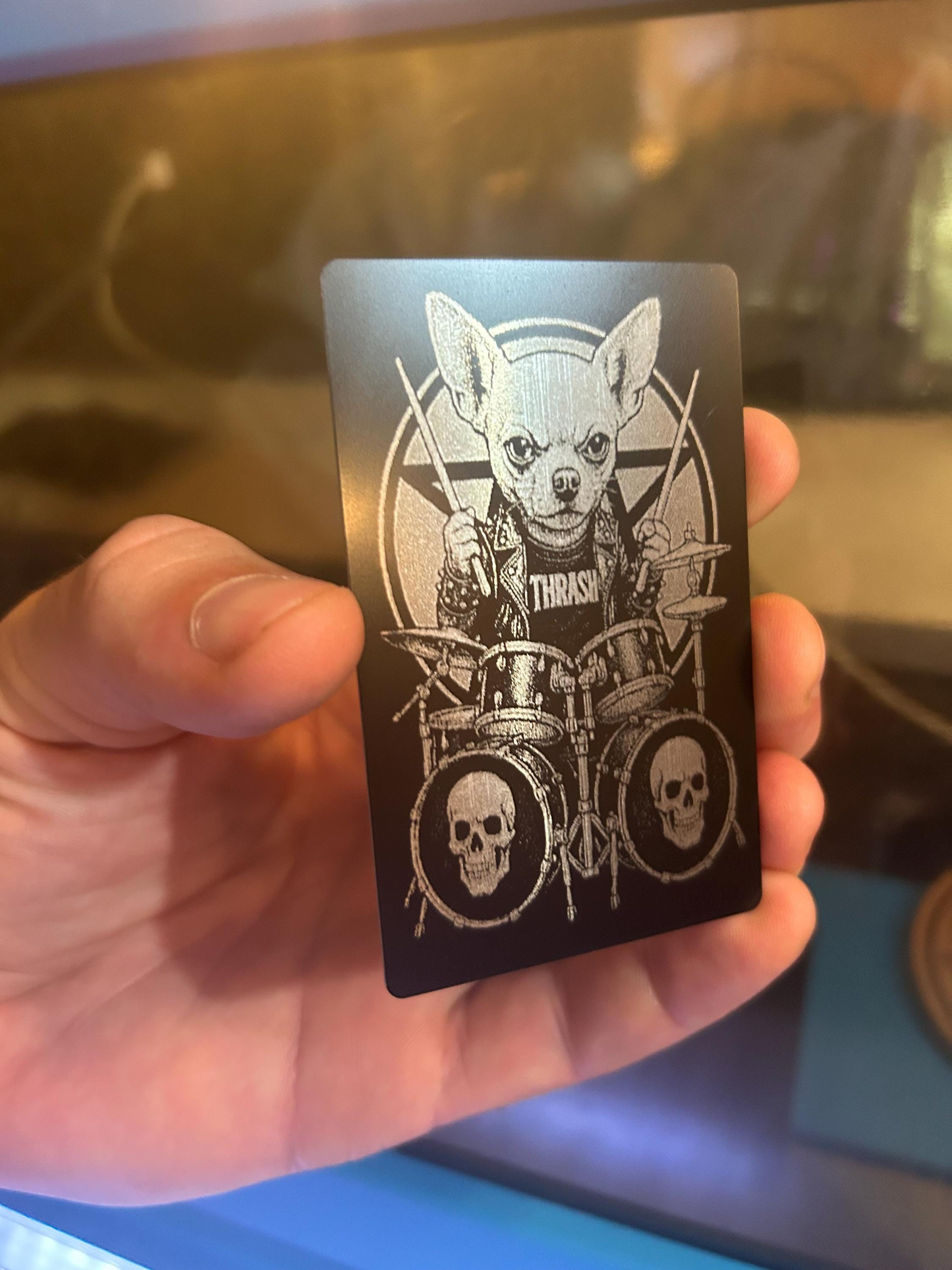 Hand holding a laser engraved metal keepsake placard with a cute funny dog metal drummer illustration and 'Thrash' text against a reflective surface.