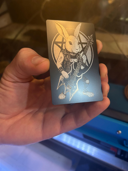 Hand holding a laser engraved metal keepsake card with black/silver illustration art of heavy metal rocker bunny with bass guitar