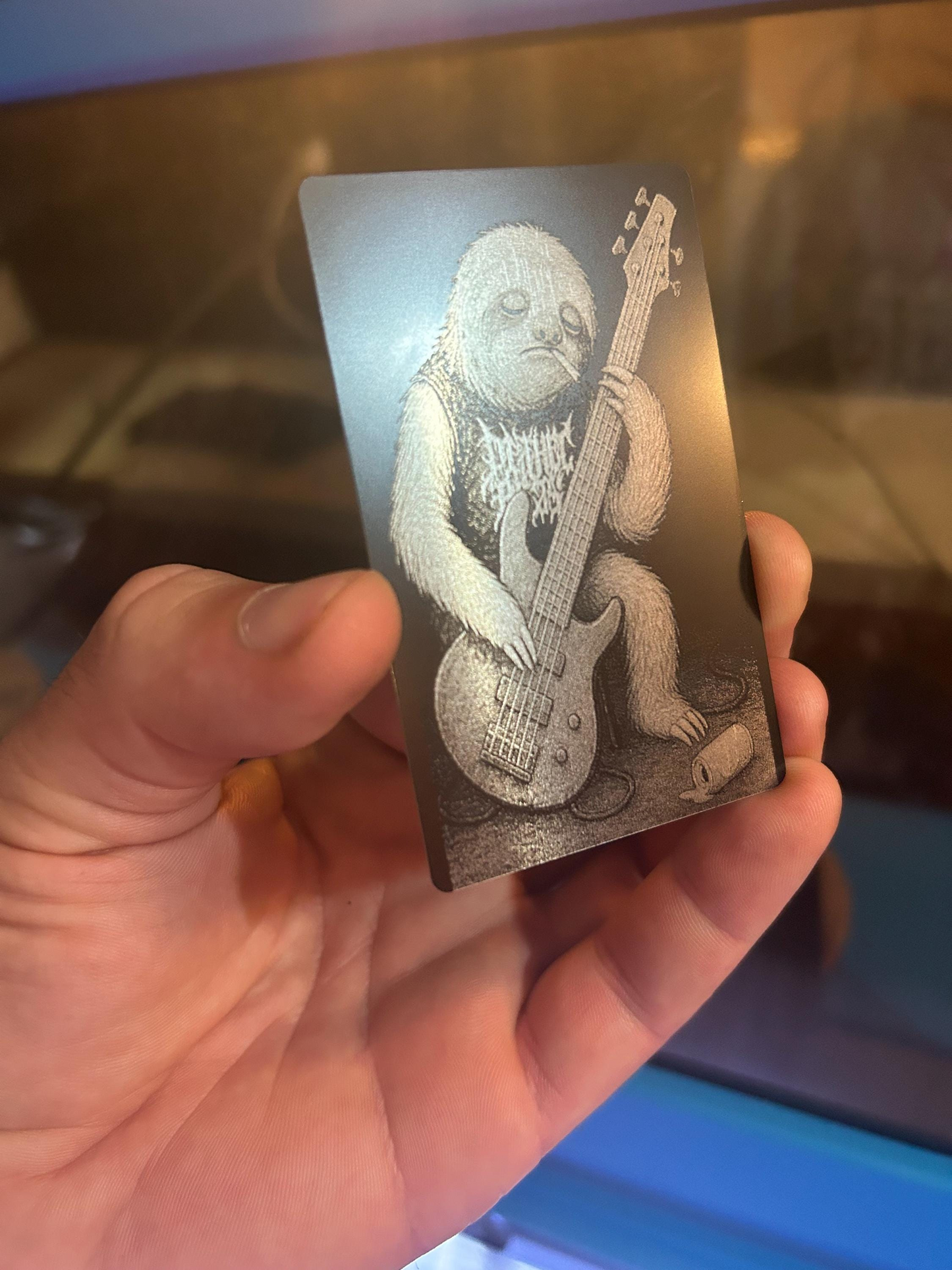 Hand holding a metal keepsake card with a engraved illustration of a hipster rockstar sloth playing an extended range bass guitar.