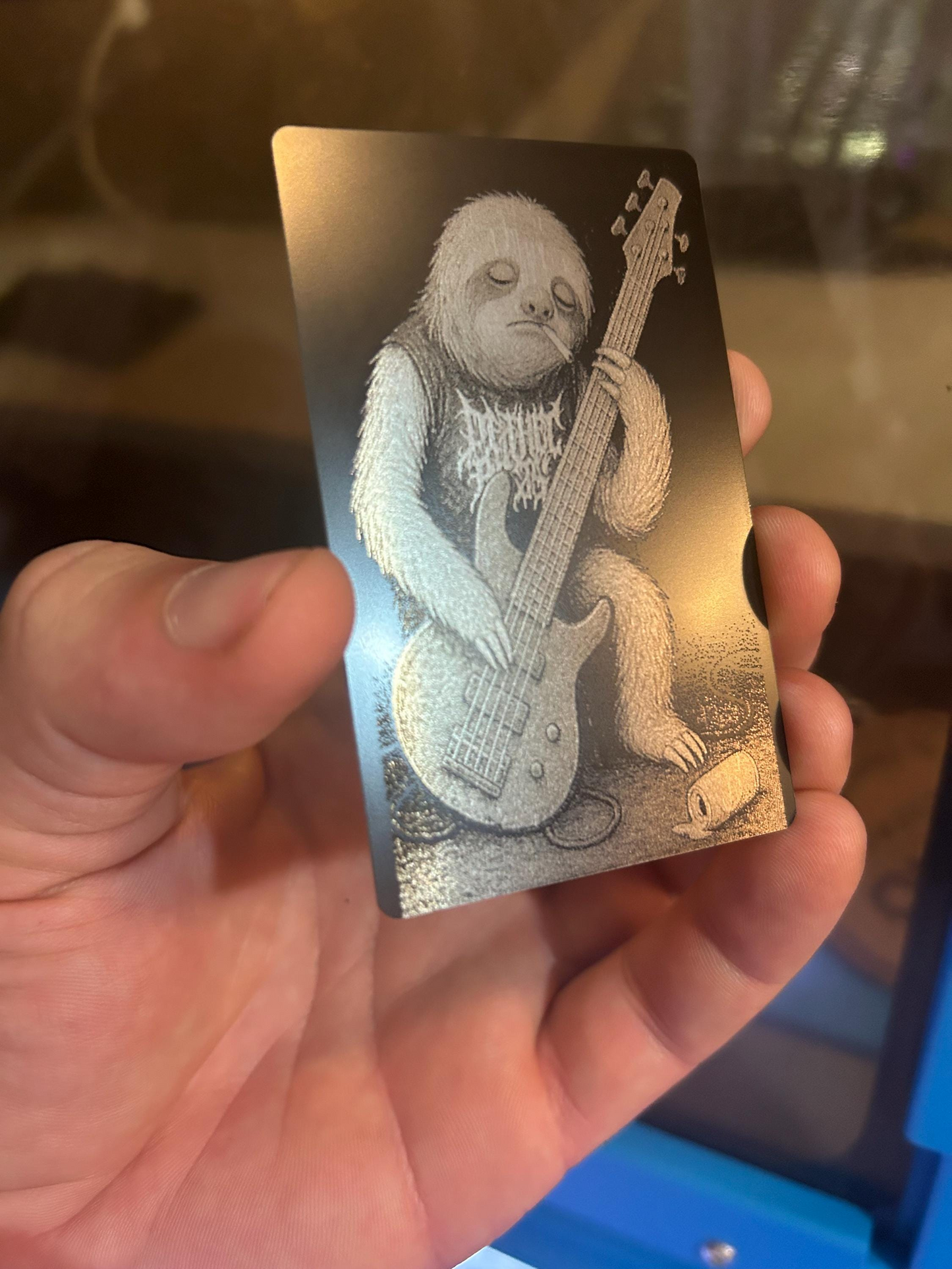 Hand holding a metal gift card with a funny cute illustration of a silly sloth playing a 5 string bass guitar.