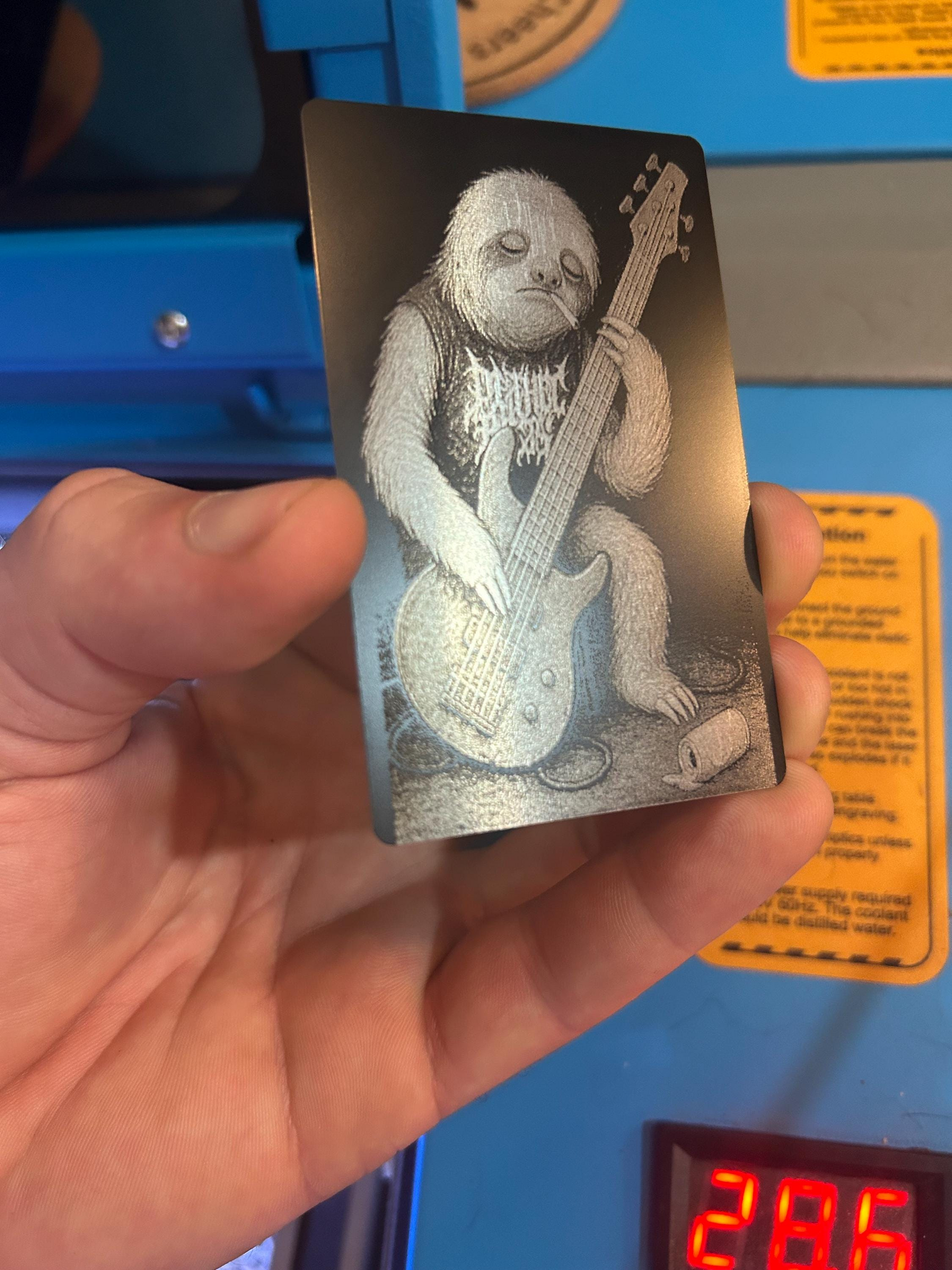 Hand holding a laser engraved anodized aluminum metal card with a design of a sloth playing a guitar against a blue background.