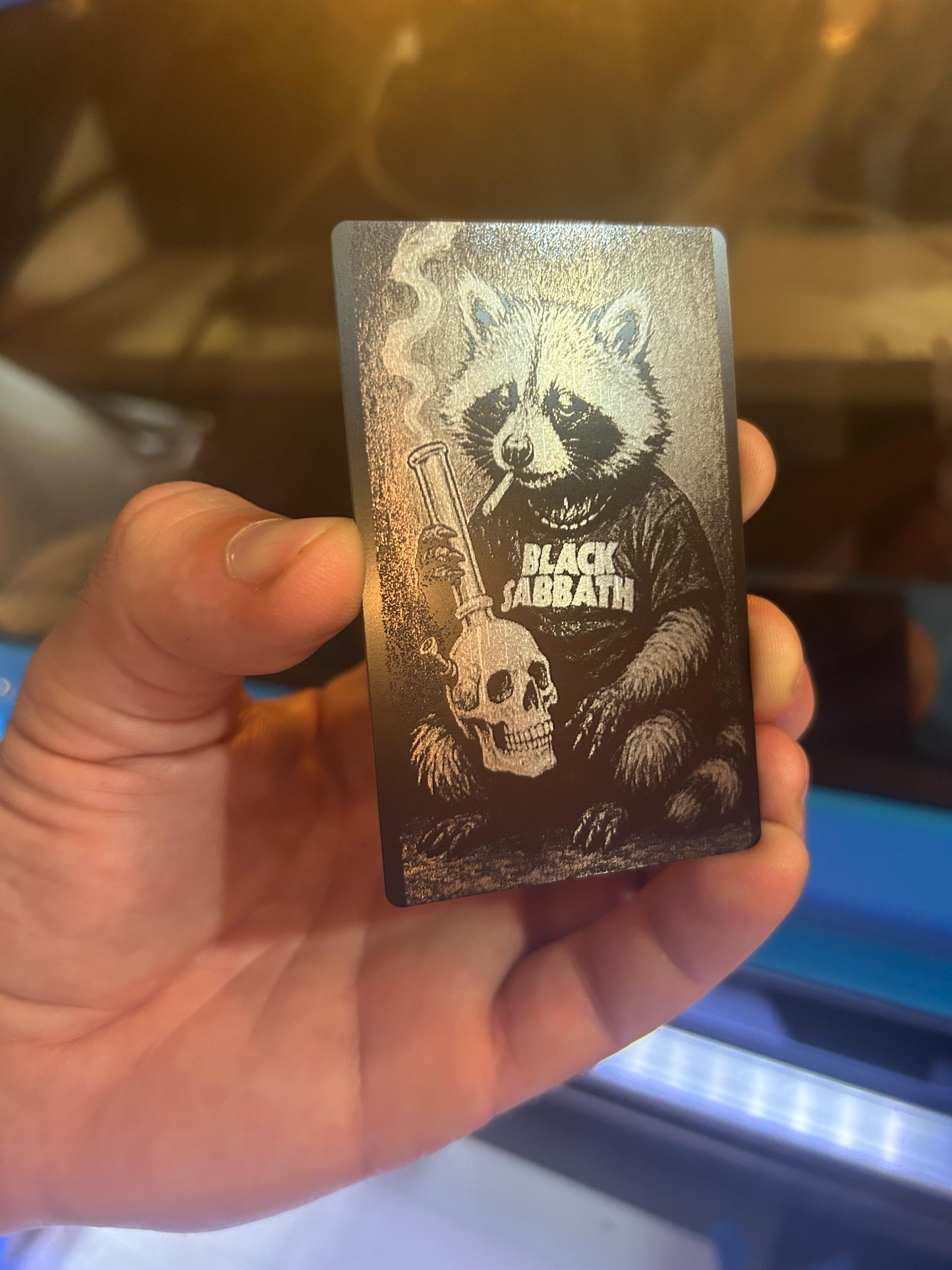 Hand holding a laser engraved black/silver metal keepsake wallet insert, with heavy metal stoner raccoon illustration design in front of a blurred machine background