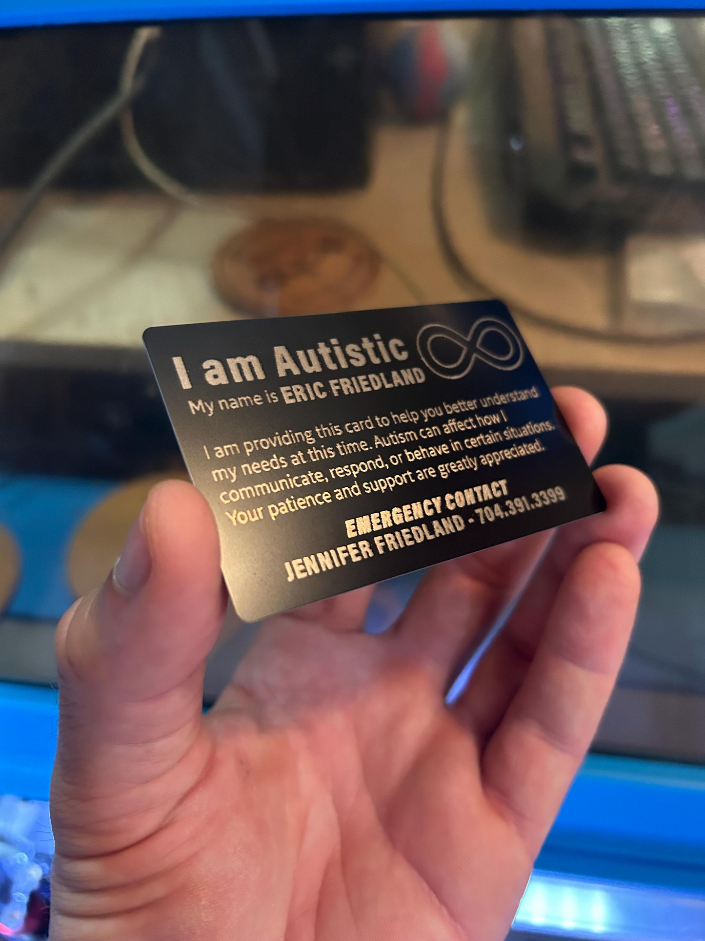 Hand holding a 'I am Autistic' identity metal business card with sample personal information on a blurred background