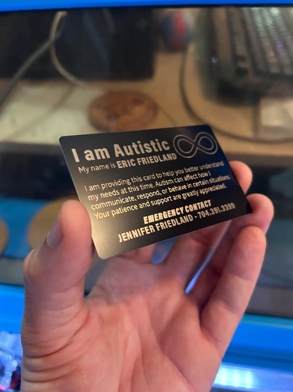 Hand holding a 'I am Autistic' identity metal business card with sample personal information on a blurred background
