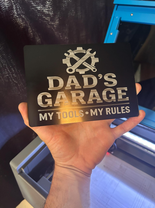 Hand holding a metal laser engraved sign with 'Dad's Garage' and 'My Tools, My Rules' text.