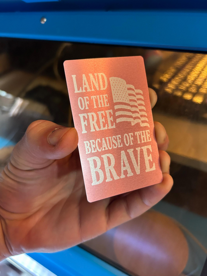 Hand holding a pink card with 'Land of the Free Because of the Brave' text and American flag design.