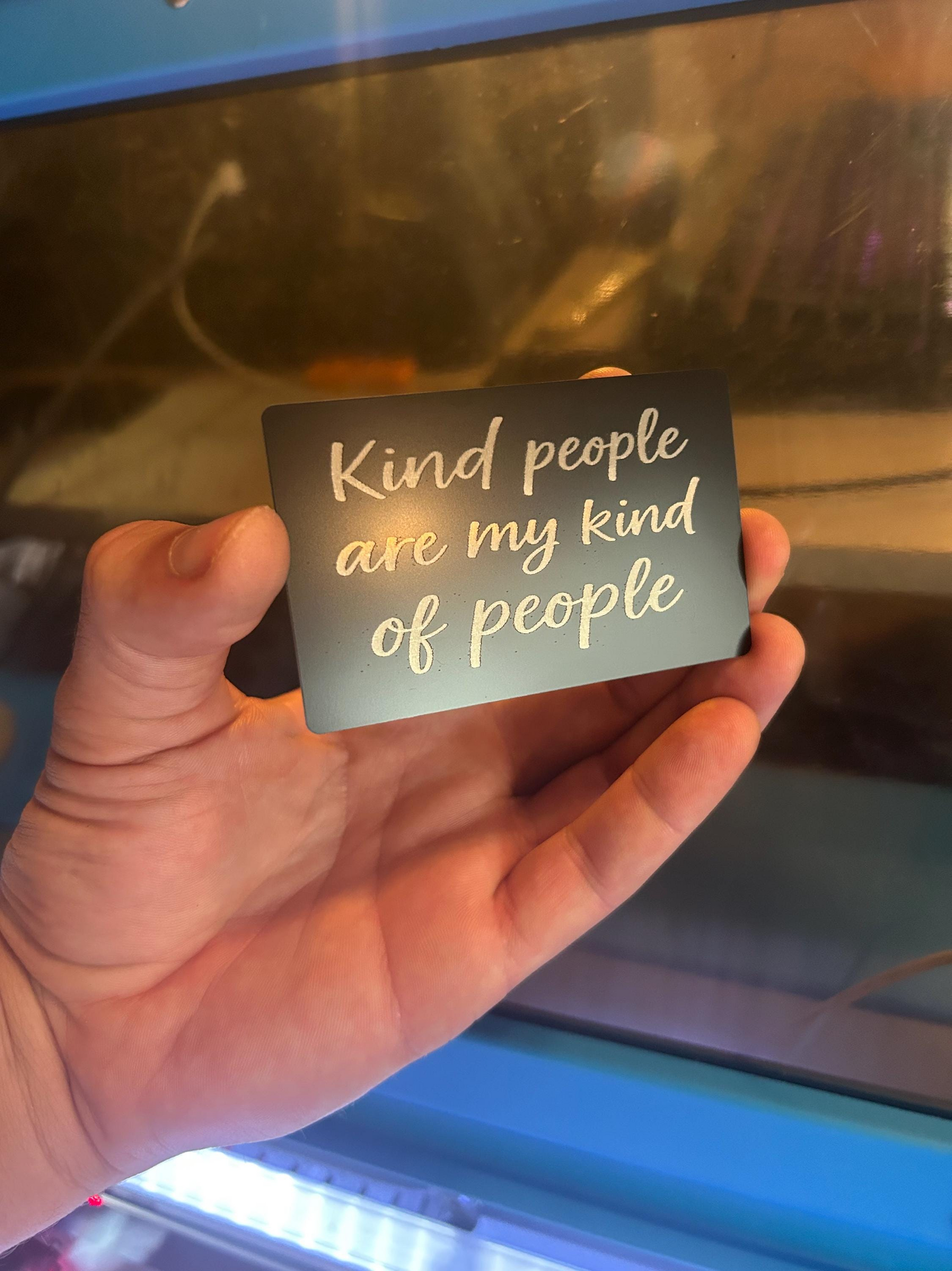 Hand holding a metal keepsake card with 'Kind people are my kind of people' text against a reflective surface.