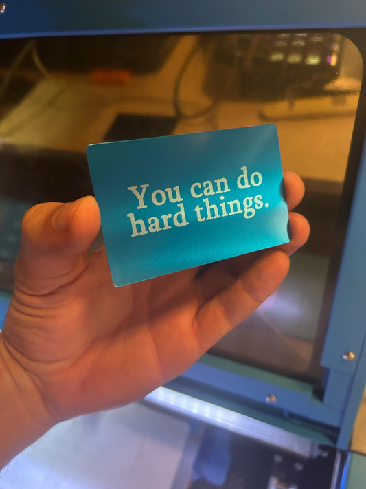 Hand holding a blue laser engraved metal gift card with 'You can do hard things.' text in front of a machine.