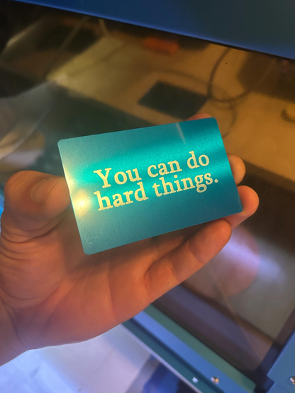 Hand holding a blue and silver metal inspirational wallet insert with 'You can do hard things.' text on a reflective surface.