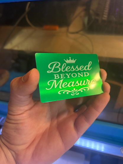 Hand holding a laser engraved metal business card with Blessed Beyond Measure keepsake card in green/silver in front of a laser engraving machine