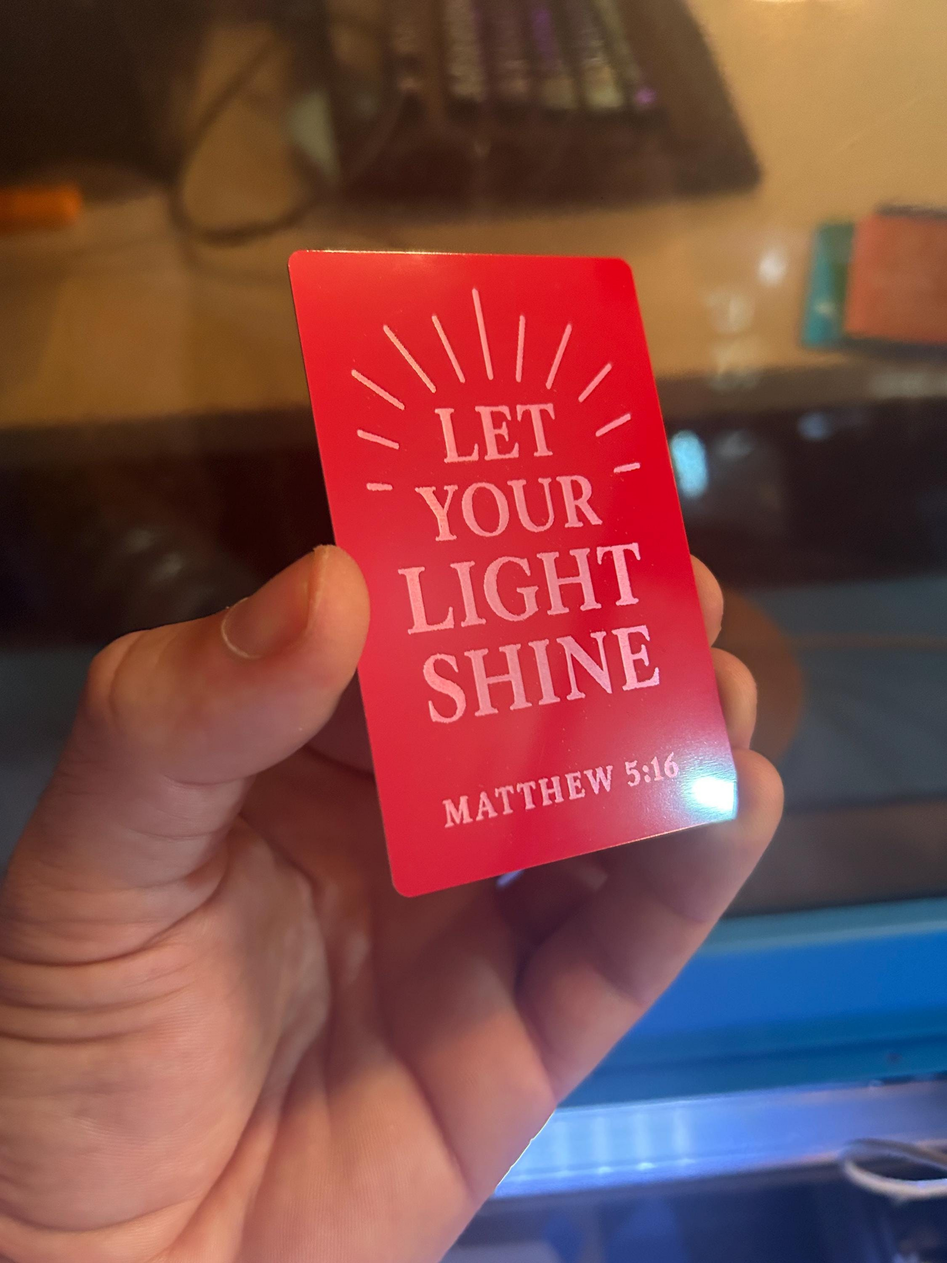 Angled shot of laser engraved red and silver card with Matthew 5:16 scripture and sunburst design held in hand