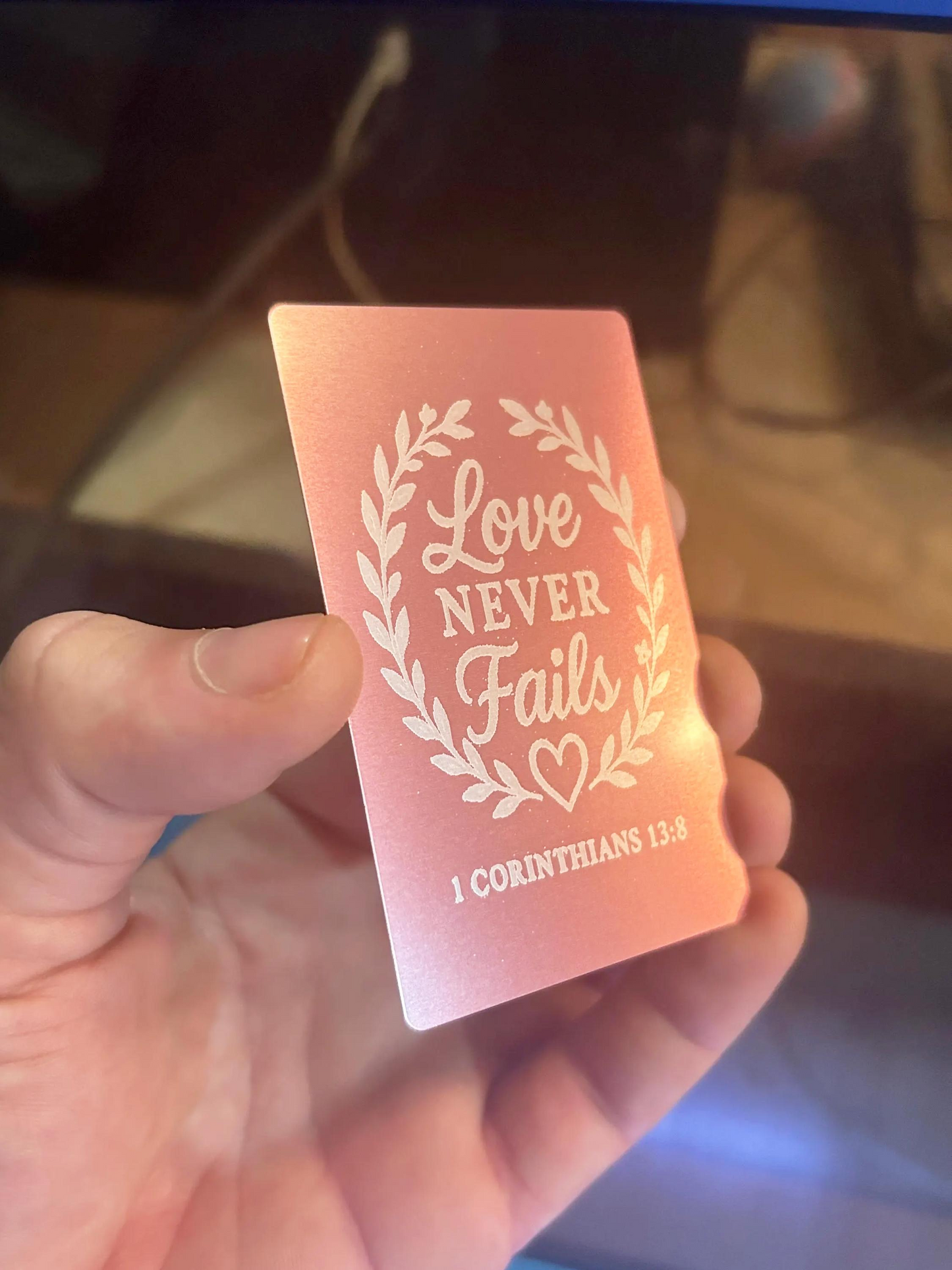 Close-up of hand displaying a metal rose gold inspirational keepsake card with Love Never Fails typography design and 1 Corinthians 13:8 bible reference below