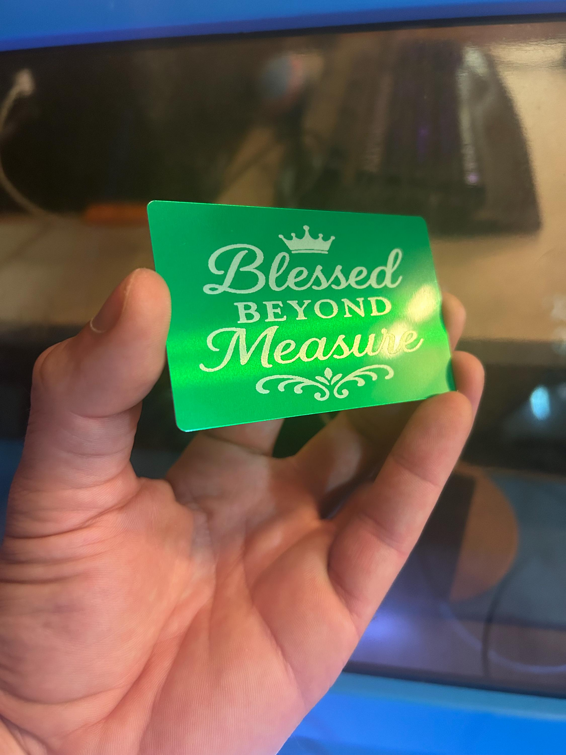 Hand holding a green/silver laser engraved card with 'Blessed Beyond Measure' text against a reflective surface.