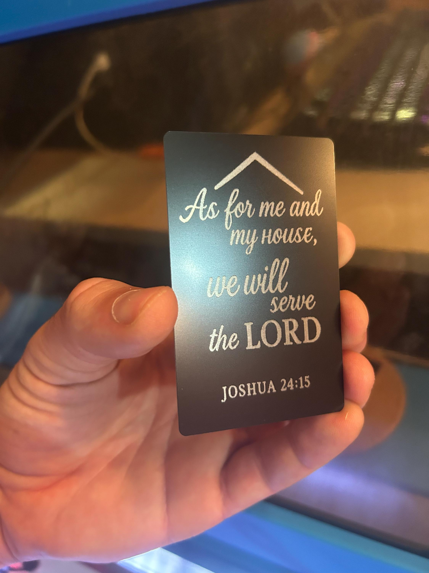 Hand holding a black metal keepsake gift card with silver text 'As for me and my house, we will serve the LORD' from Joshua 24:15.