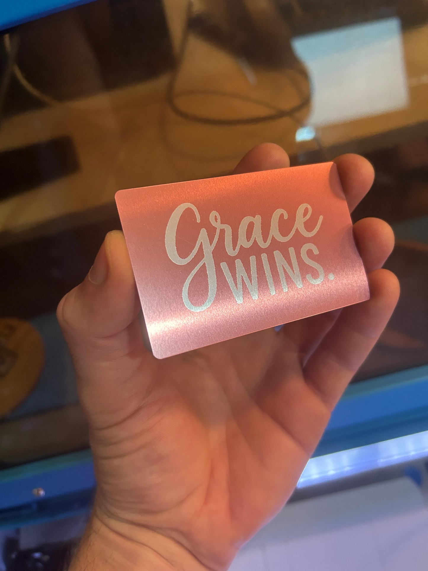 Hand holding a rose gold metal novelty card with 'Grace Wins' text against a blurred background