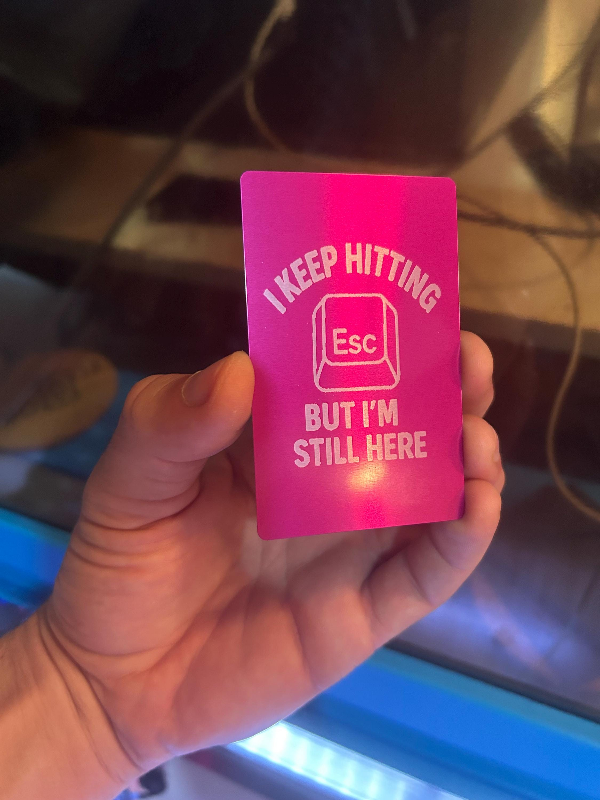 Hand holding a laser engraved metal keepsake card with "I keep hitting escape, but I'm still here" text with a computer keyboard button graphic