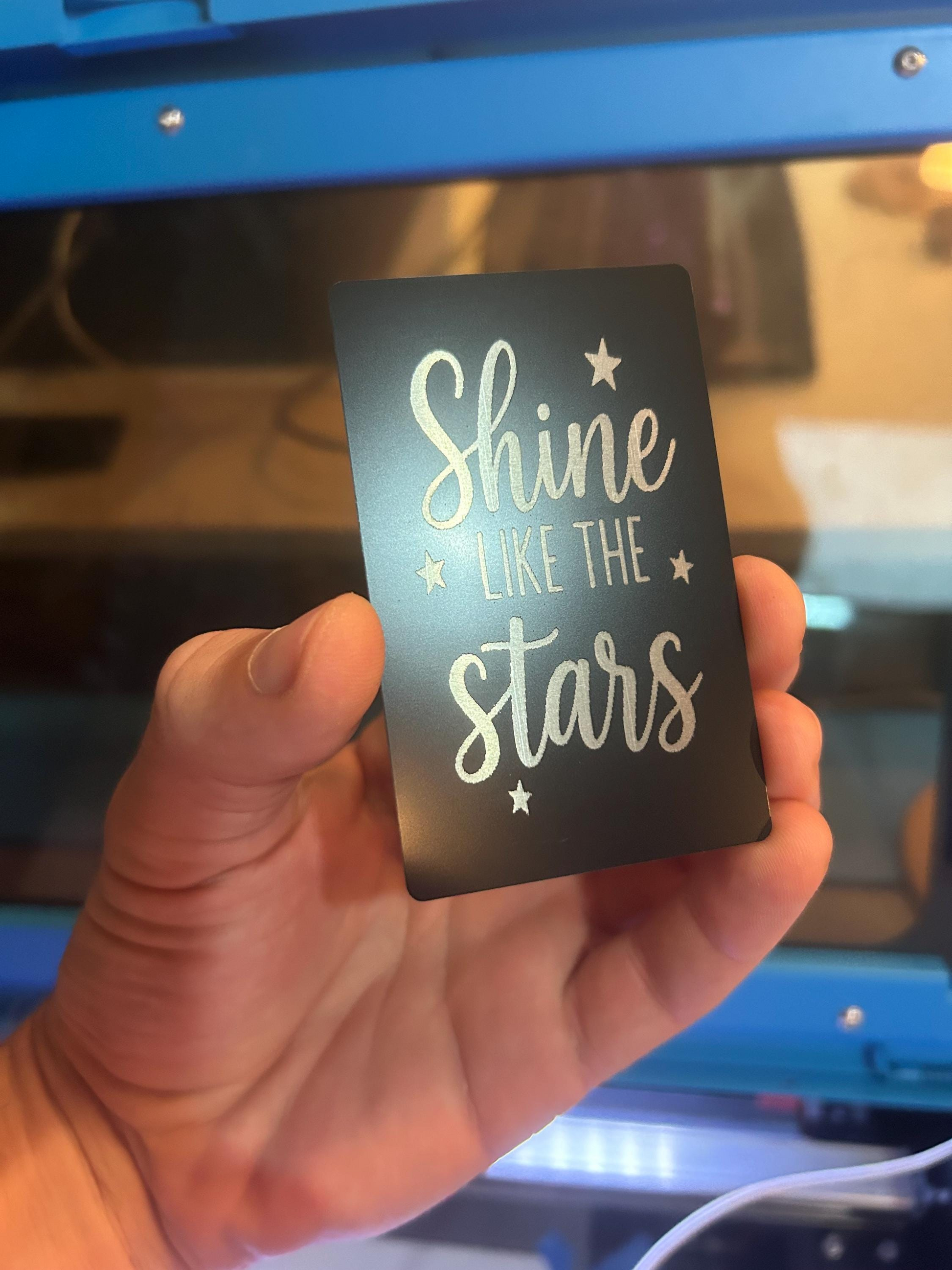 Hand holding a laser engraved "shine like the stars" metal gift card in matte black/silver