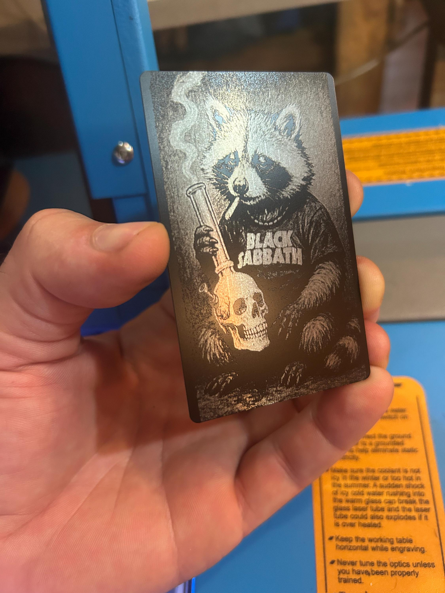 Hand holding a laser engraved metal keepsake card with black/silver doom metal raccoon design with hippie hipster cute heavy metal animal design in front of a laser engraving machine