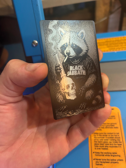 Hand holding a laser engraved metal keepsake card with black/silver doom metal raccoon design with hippie hipster cute heavy metal animal design in front of a laser engraving machine