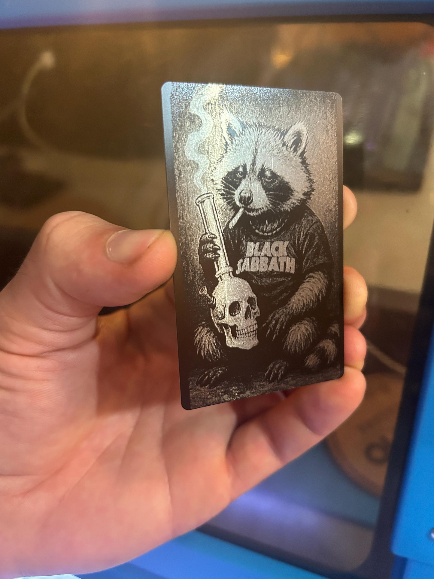 Hand holding a card with a black and silver laser engraved illustration of a raccoon and skull