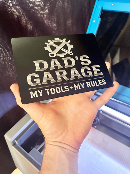 Hand holding a black/silver sign with 'Dad's Garage, My Tools, My Rules' text.