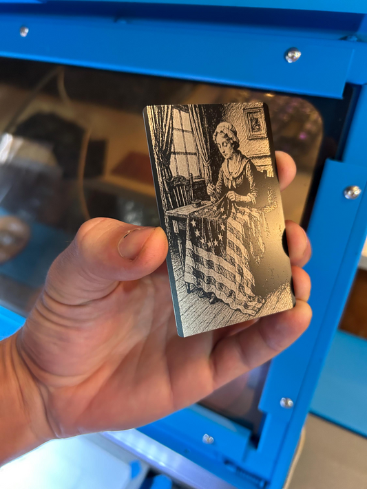 Hand holding a metal card with an engraved image of a woman in a room, against a blue background.