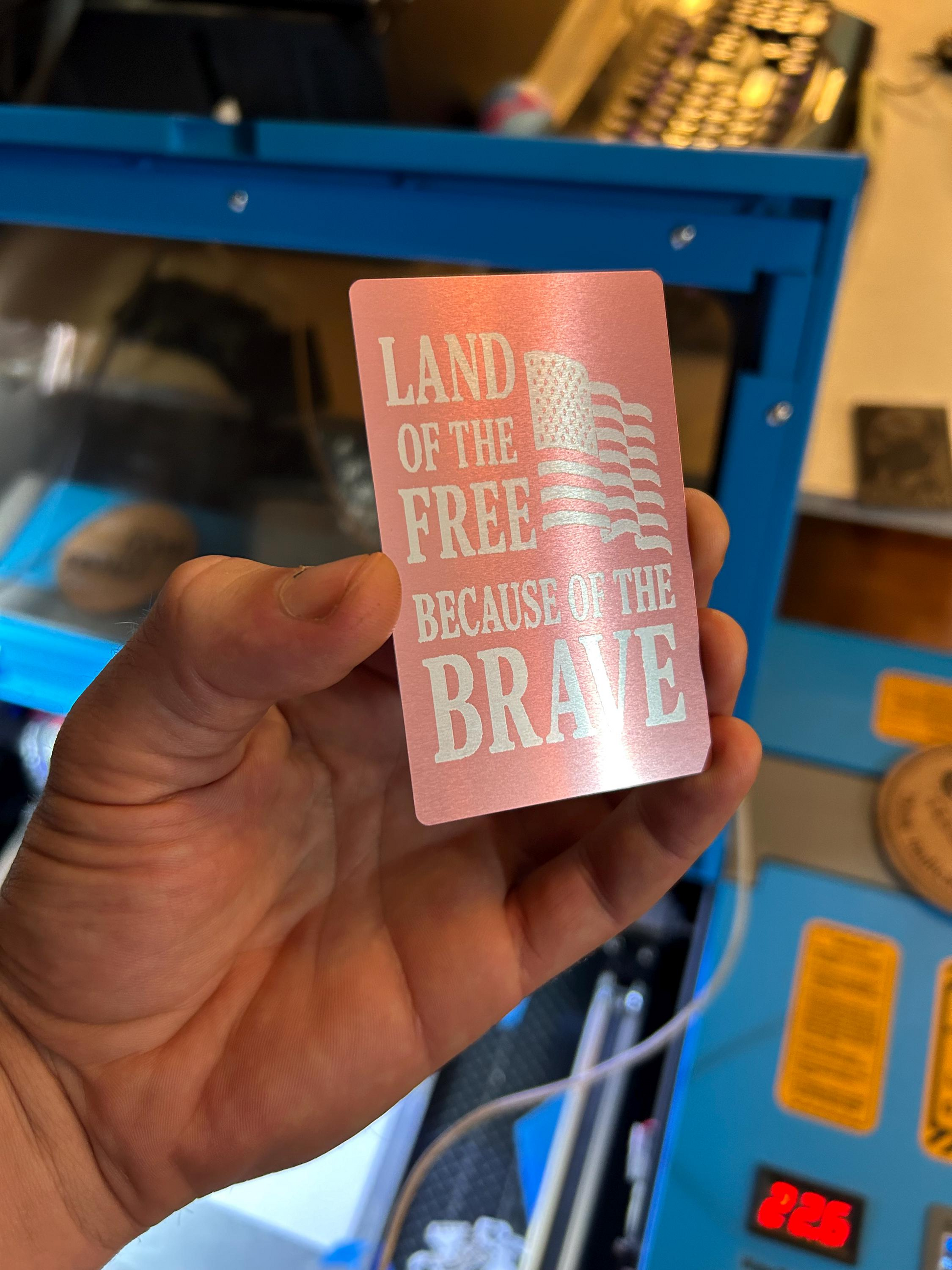 Hand holding a metal rose gold card with 'Land of the Free Because of the Brave' text and American flag design.